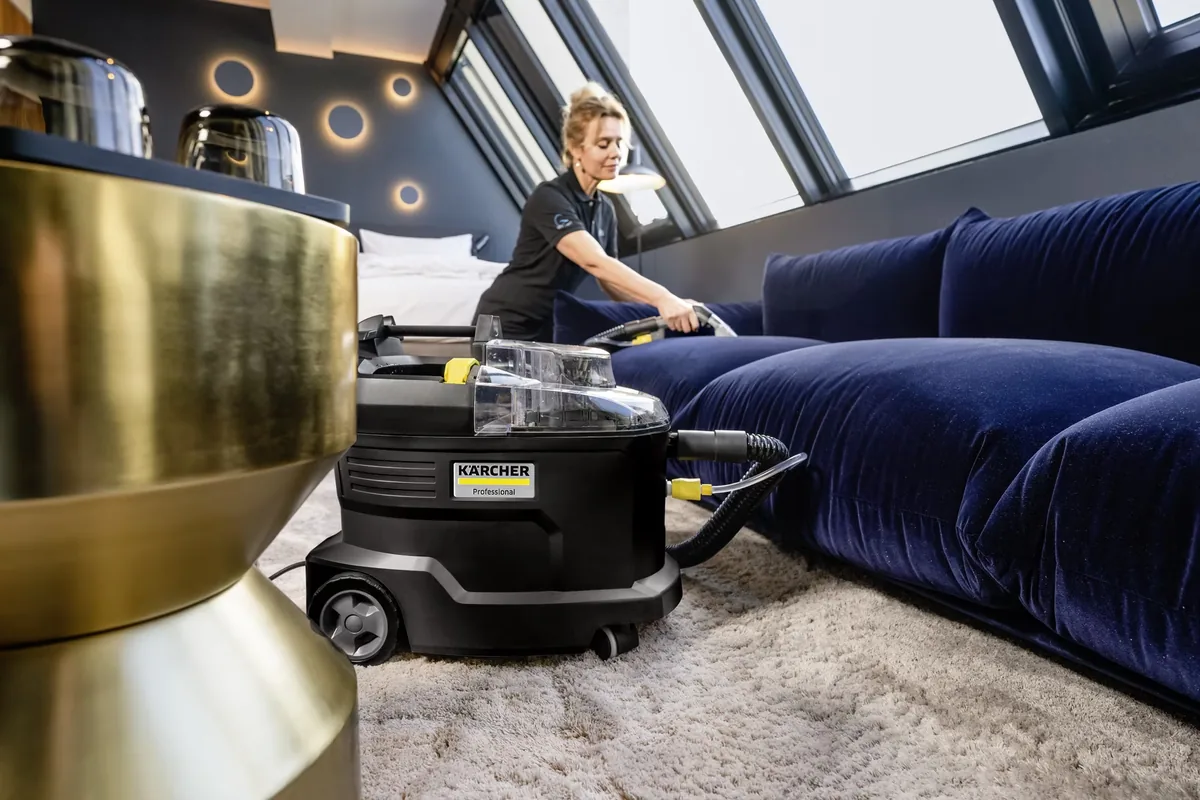 Nhân viên vệ sinh dùng máy phun hút Kärcher Professional làm sạch ghế sofa nhung xanh trong phòng khách sạn